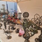 Stoisko z metalowymi rzeźbami na białym obrusie, w tym zegary i serca. Tło beżowe, namiot. Kolory srebrny, czerwony i zielony.