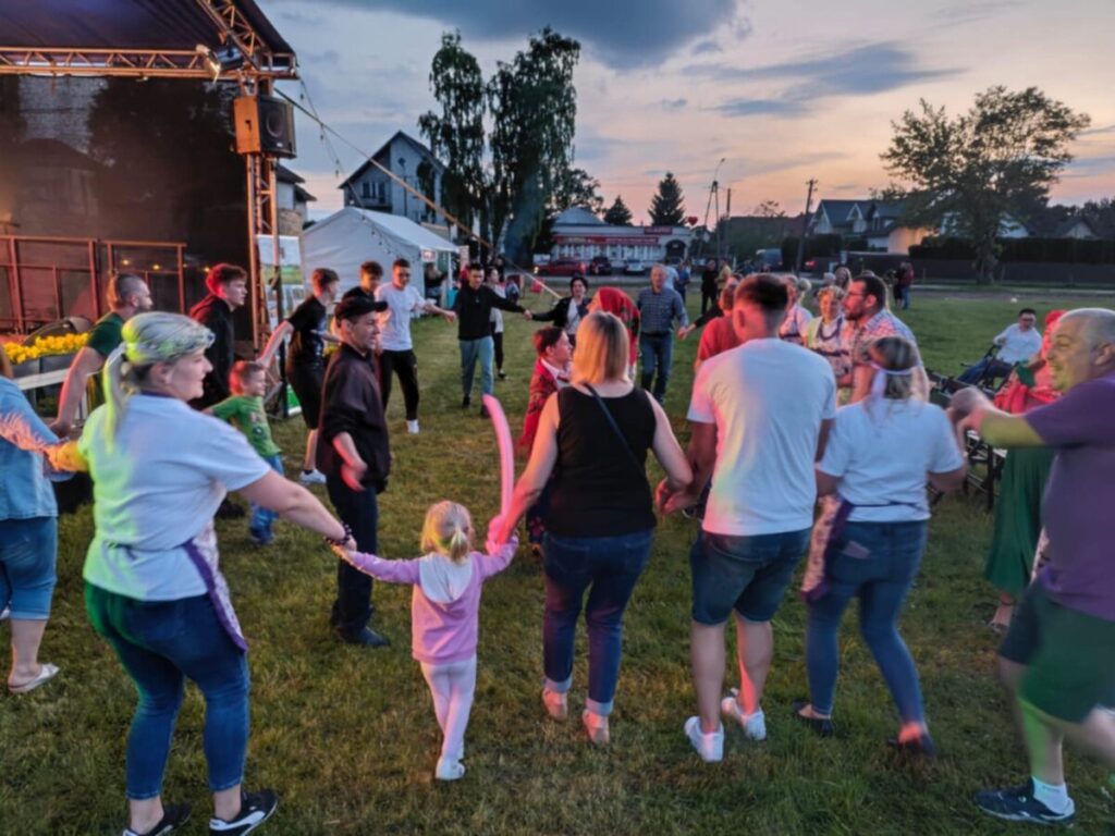Grupa ludzi tańczy w kręgu na trawniku wieczorem. Kolory stroje uczestników są różnorodne. W tle scena i namioty, a niebo jest pomarańczowo-niebieskie o zachodzie słońca.
