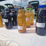 Na białym obrusie stoją słoiki z sokami domowej roboty w różnych kolorach. W tle otwarte bagażniki aut. Pogodny dzień z lekkim zachmurzeniem.