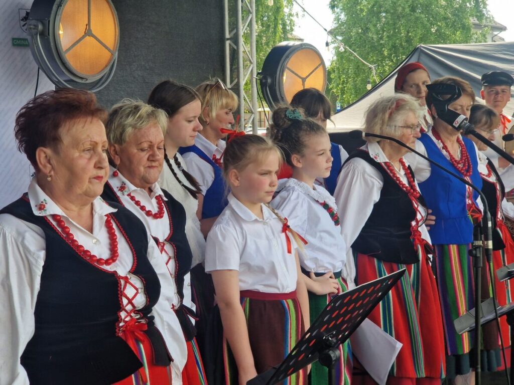 Grupa osób w tradycyjnych strojach ludowych śpiewa na scenie. Dominują kolory biały, czerwony i czarny. W tle widać zielone drzewa.