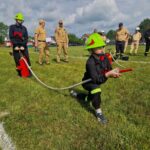 Grupa osób w strażackich uniformach bierze udział w ćwiczeniach na zielonym trawniku. Dziecko w jasnym hełmie trzyma wąż strażacki. Obserwują ich inni w beżowych mundurach na tle drzew i chmur.