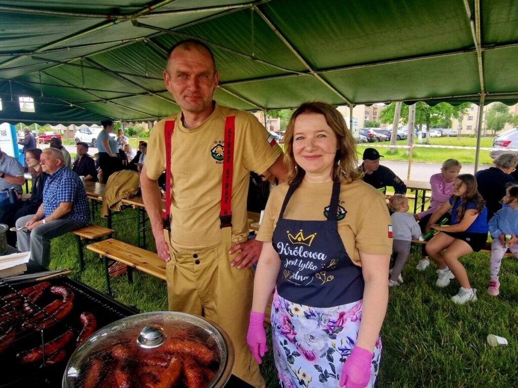 Dwóch dorosłych w jasnych ubraniach stoi pod zielonym namiotem. Na grillu pieką się kiełbaski. W tle widać siedzących ludzi i zieleń.