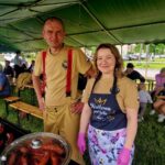 Dwóch dorosłych w jasnych ubraniach stoi pod zielonym namiotem. Na grillu pieką się kiełbaski. W tle widać siedzących ludzi i zieleń.