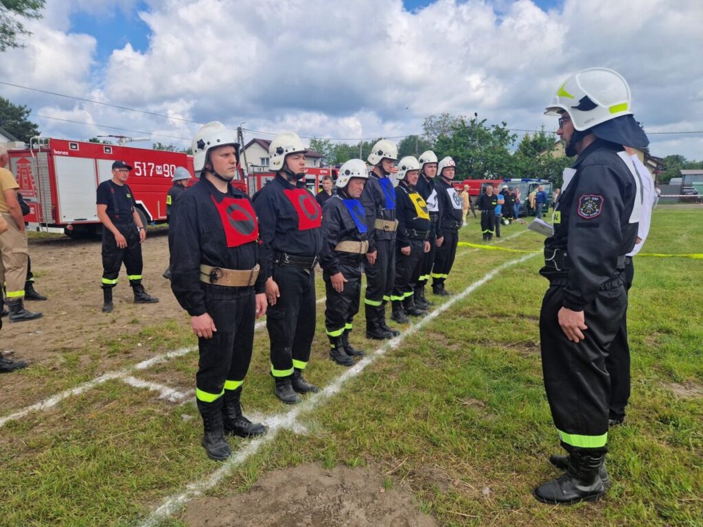 Grupa strażaków w czarnych mundurach stoi na zielonej trawie, ustawiona w szeregu, przed czerwonymi wozami strażackimi. Każdy strażak nosi biały hełm i różnokolorowe oznaczenia na klatce piersiowej. T