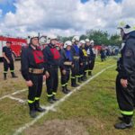 Grupa strażaków w czarnych mundurach stoi na zielonej trawie, ustawiona w szeregu, przed czerwonymi wozami strażackimi. Każdy strażak nosi biały hełm i różnokolorowe oznaczenia na klatce piersiowej. T