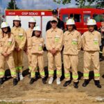 Grupa młodych strażaków w beżowych mundurach z białymi kaskami stoi przed czerwonym wozem strażackim. Za nimi niebo z chmurami. Na mundurach zielone napisy STRAŻ.