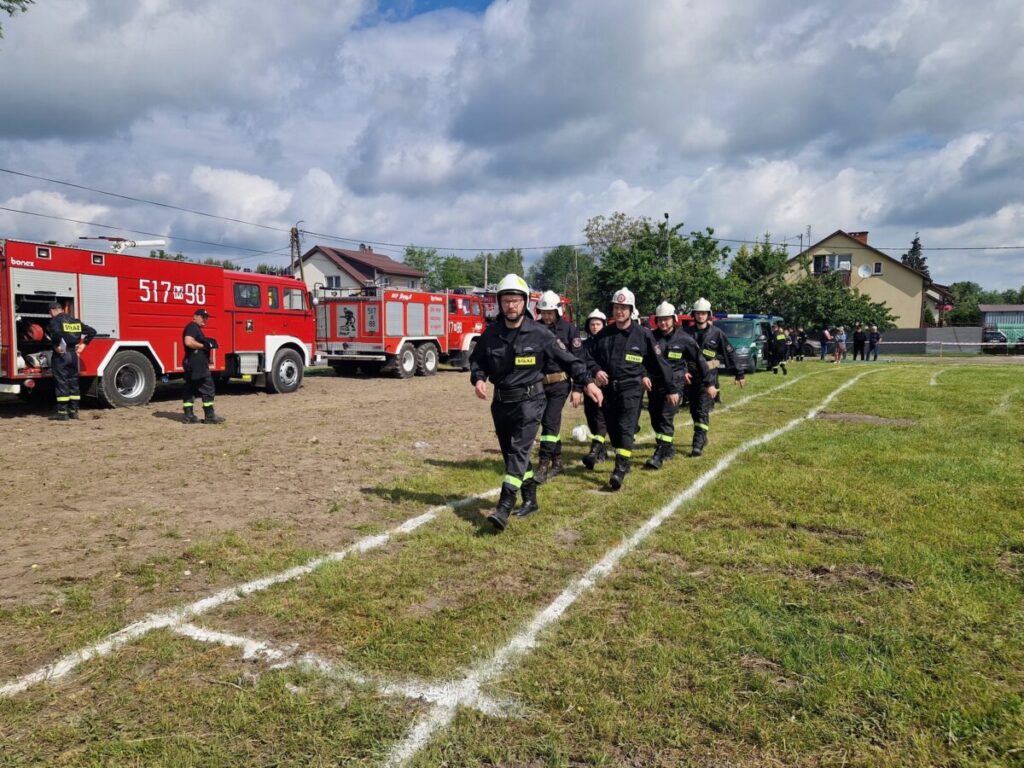 Grupa strażaków w czarnych mundurach maszeruje po trawie. W tle czerwone wozy strażackie i błękitne niebo. Obok nich kilka domów i drzewa.