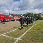 Grupa strażaków w czarnych mundurach maszeruje po trawie. W tle czerwone wozy strażackie i błękitne niebo. Obok nich kilka domów i drzewa.