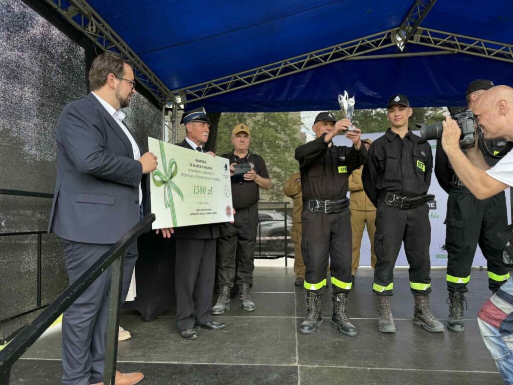 Na scenie grupa strażaków odbiera nagrodę. Mężczyzna trzyma duży czek na 1500 zł. Tło w kolorze niebieskim i czarnym.