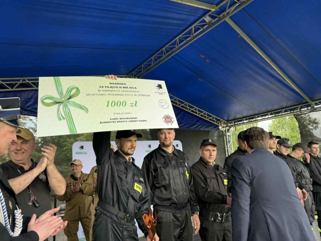 Nie wiem, kto jest na zdjęciu, ale widzę grupę ludzi w ciemnych uniformach stojących na scenie z niebieskim zadaszeniem. Jeden z nich trzyma dużą nagrodę pieniężną o wartości 1000 zł z zieloną kokardą. W tle znajdują się drzewa