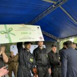 Nie wiem, kto jest na zdjęciu, ale widzę grupę ludzi w ciemnych uniformach stojących na scenie z niebieskim zadaszeniem. Jeden z nich trzyma dużą nagrodę pieniężną o wartości 1000 zł z zieloną kokardą. W tle znajdują się drzewa