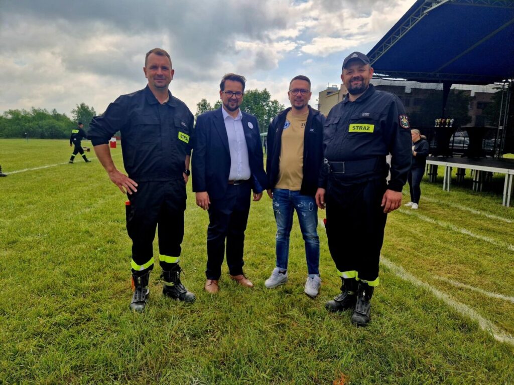 Grupa czterech mężczyzn stoi na zielonej trawie. Dwóch nosi czarne mundury strażackie. W tle widać scenę i drzewa pod pochmurnym niebem.