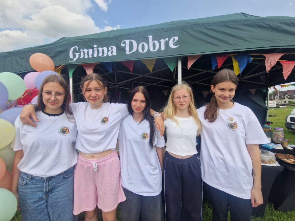 Grupa pięciu młodych osób w białych koszulkach stoi przed zielonym namiotem z napisem Gmina Dobre. Tło zdobią kolorowe balony i girlandy. Namiot jest ustawiony na trawie podczas wydarzenia na świeżym powietrzu.