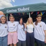 Grupa pięciu młodych osób w białych koszulkach stoi przed zielonym namiotem z napisem Gmina Dobre. Tło zdobią kolorowe balony i girlandy. Namiot jest ustawiony na trawie podczas wydarzenia na świeżym powietrzu.