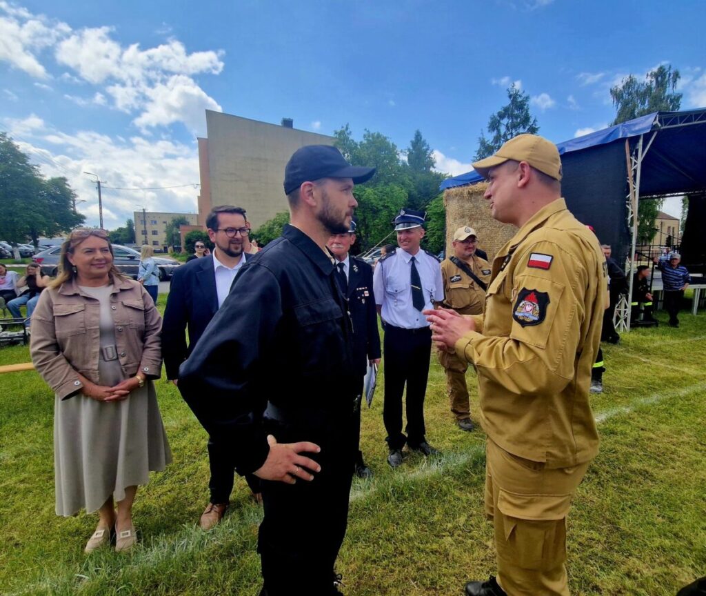 Grupa osób na otwartym terenie rozmawia w słoneczny dzień. Mężczyzna w beżowym mundurze stoi naprzeciw osoby w czerni. W tle są zielone drzewa i niebieskie niebo.