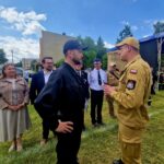 Grupa osób na otwartym terenie rozmawia w słoneczny dzień. Mężczyzna w beżowym mundurze stoi naprzeciw osoby w czerni. W tle są zielone drzewa i niebieskie niebo.