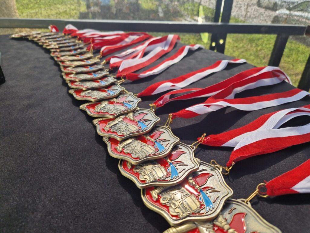 Na czarnym tle leży rząd złotych medali z biało-czerwonymi wstążkami. Medale mają czerwono-niebieskie detale i wzór nawiązujący do strażaków. W tle widać zieloną trawę przez siatkę.