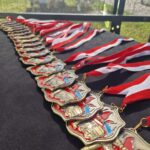 Na czarnym tle leży rząd złotych medali z biało-czerwonymi wstążkami. Medale mają czerwono-niebieskie detale i wzór nawiązujący do strażaków. W tle widać zieloną trawę przez siatkę.