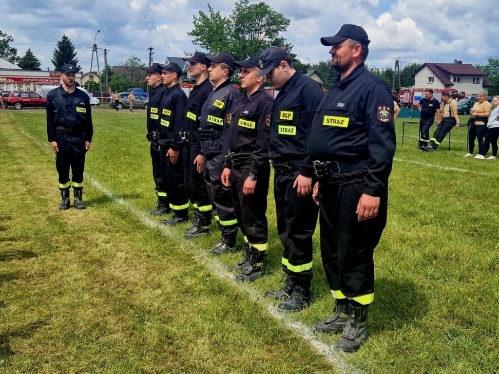 Strażacy w czarnych mundurach stoją w szeregu na zielonym trawiastym boisku. Na mundurach żółte napisy STRAŻ. W tle niebo z chmurami i kilka budynków.