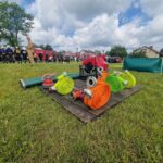 Strażacy stoją na trawie obok czerwonych wozów strażackich. Na pierwszym planie kolorowe węże strażackie na drewnianej platformie. W tle widoczne zielone drzewa i niebo z białymi chmurami.