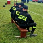 Trzech strażaków w czarnych mundurach siedzi tyłem na ławce. Na plecach mają żółte napisy STRAŻ. Tło to zielona trawa i sprzęt strażacki.