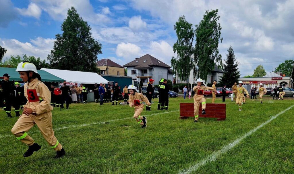 Młodzi strażacy w beżowych strojach i kaskach biegną po trawie. Tło to drzewa i namioty, pogoda pochmurna. Obok skaczą przez przeszkody.