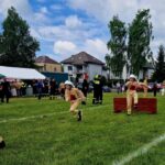 Młodzi strażacy w beżowych strojach i kaskach biegną po trawie. Tło to drzewa i namioty, pogoda pochmurna. Obok skaczą przez przeszkody.