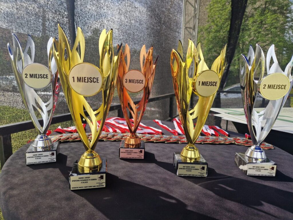 Na czarnym stole stoją trofea: złote, srebrne i brązowe, oznaczone jako 1, 2, 3 miejsce. Z tyłu widać medale. Tło jest lekko rozmyte, z fragmentem zieleni.