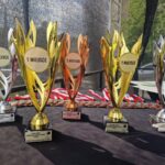 Na czarnym stole stoją trofea: złote, srebrne i brązowe, oznaczone jako 1, 2, 3 miejsce. Z tyłu widać medale. Tło jest lekko rozmyte, z fragmentem zieleni.