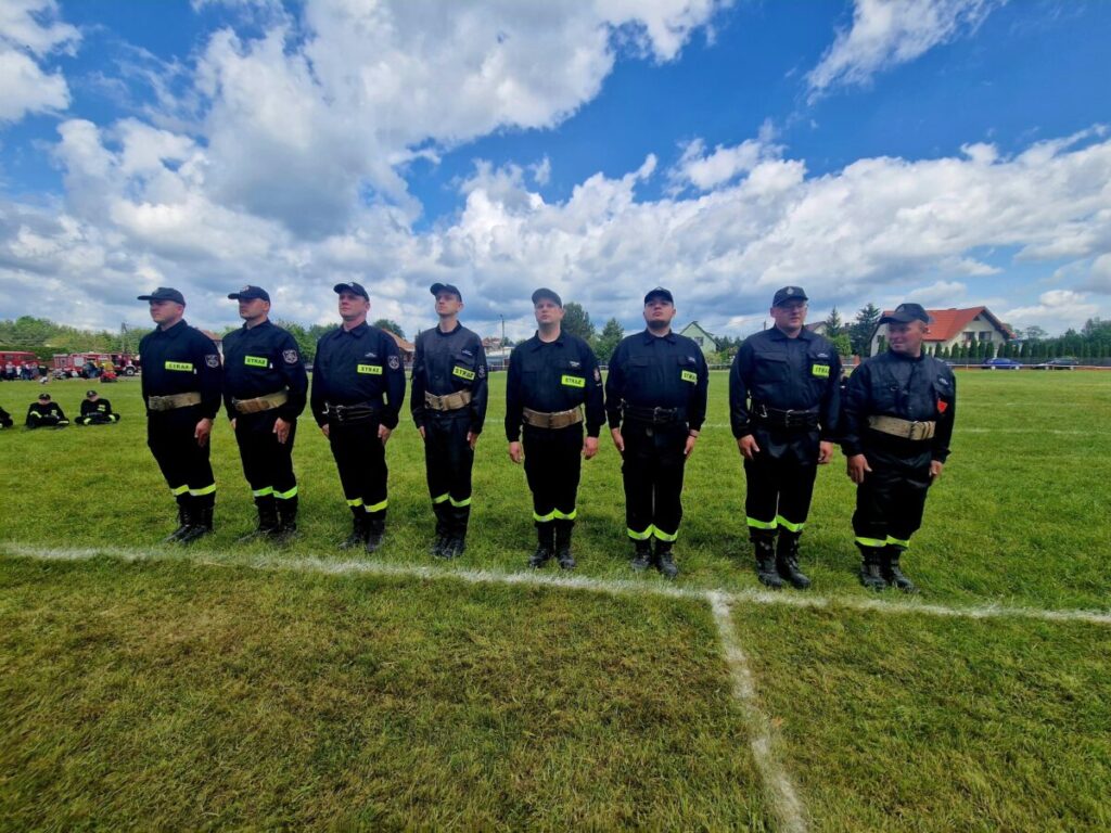 Grupa strażaków w czarnych mundurach stoi na zielonej trawie. Niebo jest jasne z białymi chmurami. W tle widać dom i pojazdy.