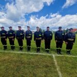 Grupa strażaków w czarnych mundurach stoi na zielonej trawie. Niebo jest jasne z białymi chmurami. W tle widać dom i pojazdy.