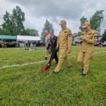 Grupa osób w beżowych mundurach chodzi po trawie z narzędziem pomiarowym. W tle namiot i drzewa. Szeroka, zielona przestrzeń.