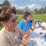 Osoba trzyma ozdobiony kamień z białym kotem, tło to zielona trawa i domy. Na stole widać przybory do malowania.