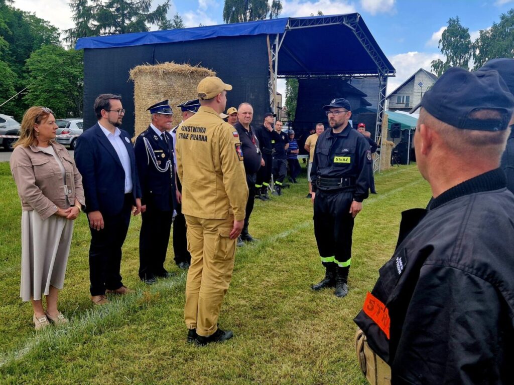 Grupa ludzi w uniformach stoi na trawie. W tle namiot i stóg siana. Jasny dzień, zielone drzewa w oddali.