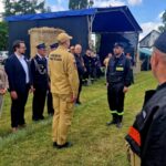 Grupa ludzi w uniformach stoi na trawie. W tle namiot i stóg siana. Jasny dzień, zielone drzewa w oddali.