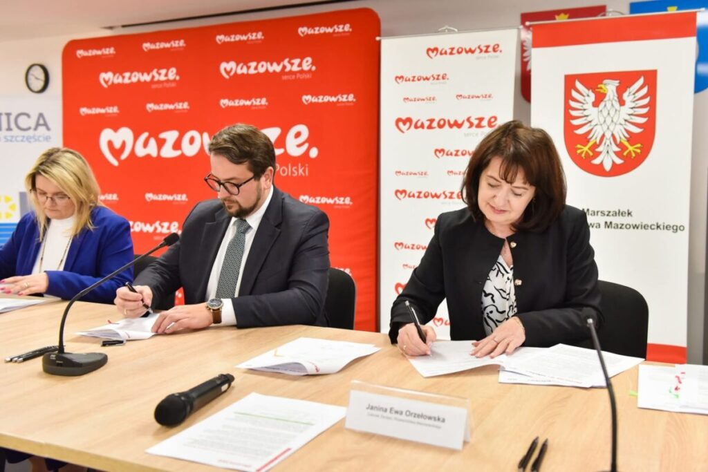 Trzy osoby podpisują dokumenty w sali z czerwonym banerem Mazowsze. Kobieta w niebieskiej marynarce, mężczyzna w garniturze i kobieta w czarnej marynarce siedzą przy drewnianym stole. Obok stoją mikrofony i tabliczka z napisem