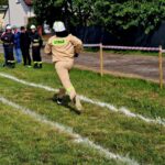 Osoba w stroju strażackim biegnie po trawie. Na lewo kilku strażaków w czarnych mundurach. W tle drzewa i ogrodzenie.