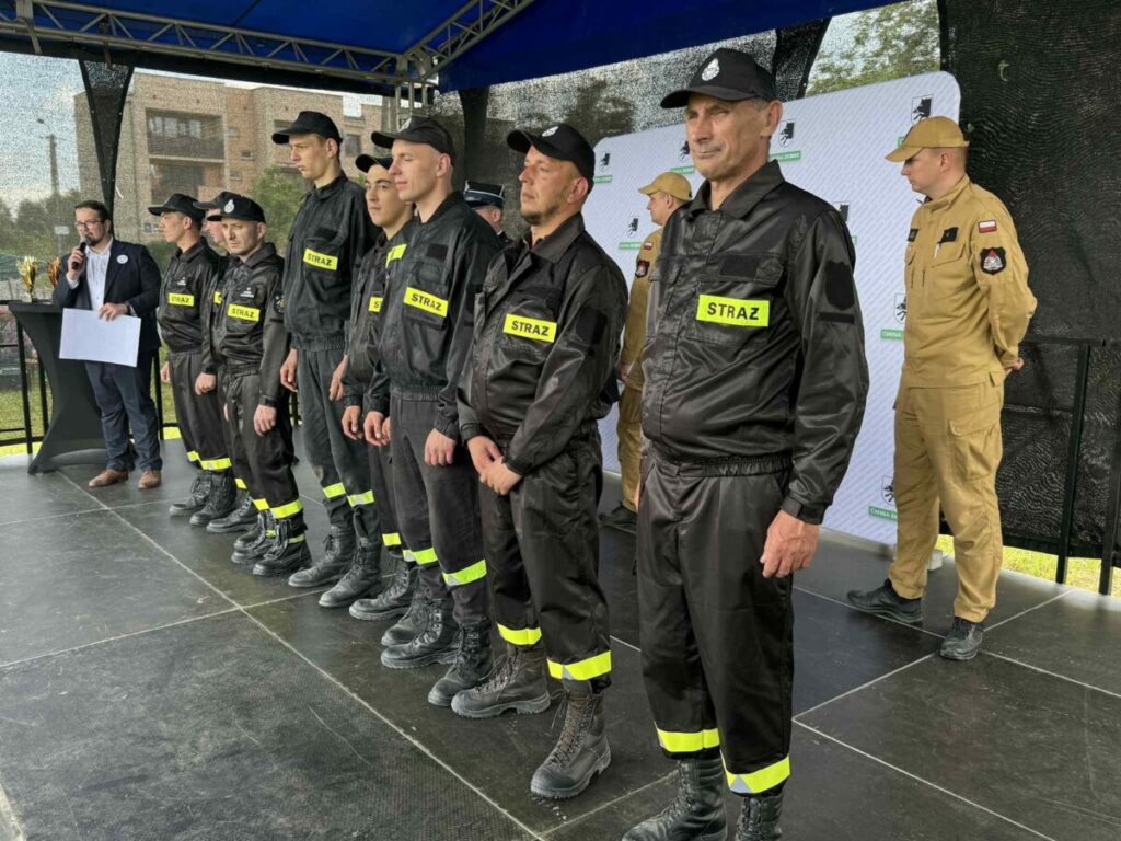 Grupa strażaków w czarnych mundurach z żółtymi napisami STRAŻ stoi na scenie pod niebieskim zadaszeniem. W tle widoczny jest szary baner i osoba przemawiająca. Scena znajduje się na świeżym powietrzu.