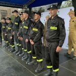 Grupa strażaków w czarnych mundurach z żółtymi napisami STRAŻ stoi na scenie pod niebieskim zadaszeniem. W tle widoczny jest szary baner i osoba przemawiająca. Scena znajduje się na świeżym powietrzu.