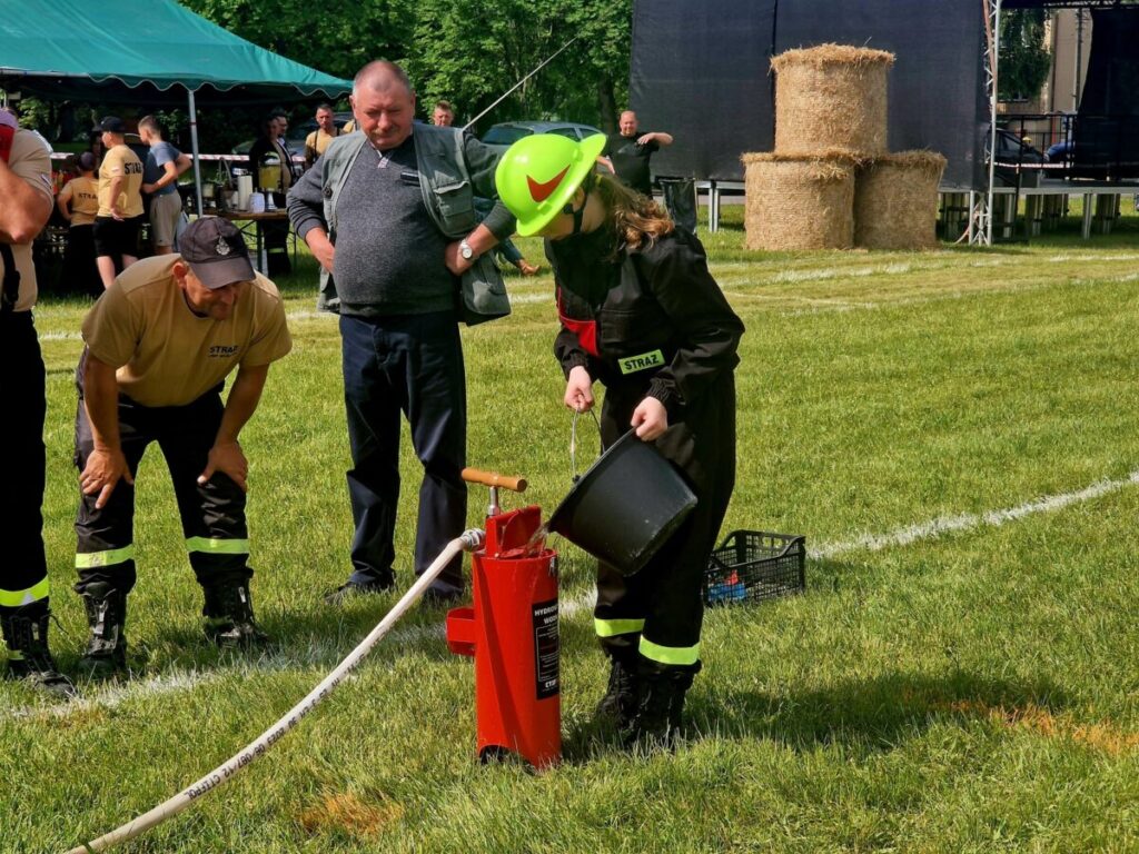 Osoba w strażackim stroju wylewa wodę z wiadra do hydrantu na trawie. Obok obserwują inni strażacy w beżowych koszulkach. W tle namiot i bele siana.