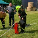 Osoba w strażackim stroju wylewa wodę z wiadra do hydrantu na trawie. Obok obserwują inni strażacy w beżowych koszulkach. W tle namiot i bele siana.