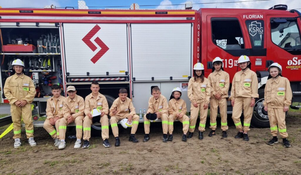 Grupa młodzieży w mundurach strażackich stoi przed czerwonym wozem strażackim. W tle otwarte wyposażenie pojazdu. Kolory: beżowy, czerwony, zielony.