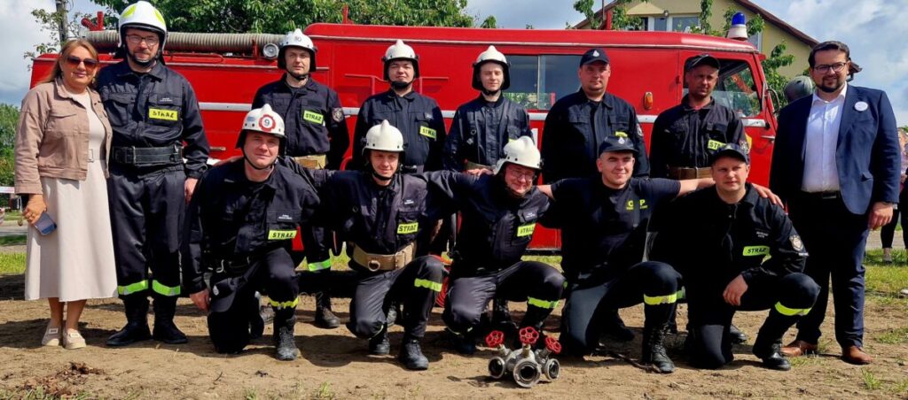 Grupa strażaków w czarnych mundurach stoi przed czerwonym wozem strażackim. W tle zielone drzewa. Obok stoi kobieta i mężczyzna w garniturze.