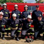 Grupa strażaków w czarnych mundurach stoi przed czerwonym wozem strażackim. W tle zielone drzewa. Obok stoi kobieta i mężczyzna w garniturze.