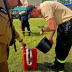 Dwóch mężczyzn w uniformach strażackich nalewa wodę z czarnego wiadra do czerwonego pojemnika. Tło to trawiaste pole z niskim budynkiem i drzewami. Słońce oświetla scenę, dając jasne, żywe kolory.