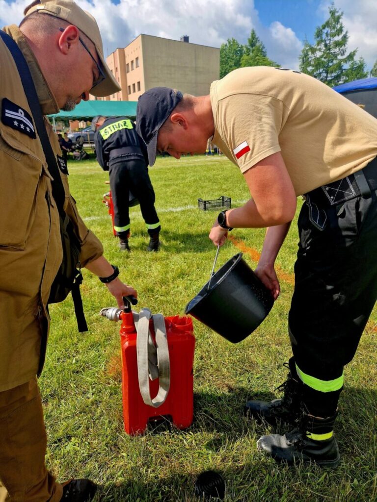 Dwóch mężczyzn w uniformach strażackich nalewa wodę z czarnego wiadra do czerwonego pojemnika. Tło to trawiaste pole z niskim budynkiem i drzewami. Słońce oświetla scenę, dając jasne, żywe kolory.