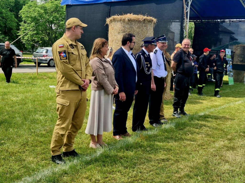 Grupa ludzi stoi w szeregu na zielonym trawniku. Mają na sobie różne mundury i ubrania formalne. W tle są bele siana i niebieski namiot.