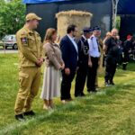 Grupa ludzi stoi w szeregu na zielonym trawniku. Mają na sobie różne mundury i ubrania formalne. W tle są bele siana i niebieski namiot.