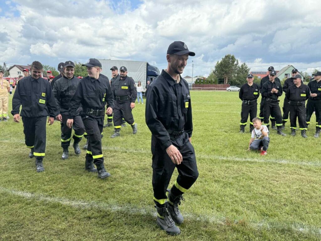 Grupa strażaków w czarnych mundurach z żółtymi elementami stoi na trawie. Jeden chłopiec siedzi na ziemi. W tle niebo z chmurami i kilka budynków.