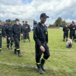 Grupa strażaków w czarnych mundurach z żółtymi elementami stoi na trawie. Jeden chłopiec siedzi na ziemi. W tle niebo z chmurami i kilka budynków.
